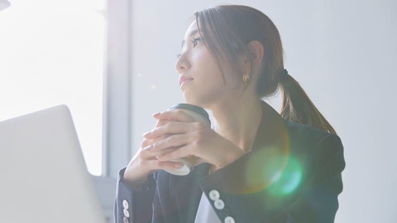 窓際でカップを持ちながら一人で悩む女性の写真で、帰宅時間が遅くなったりスマートフォンを隠すようになったりといった浮気の兆候に不安を抱え、夫婦関係の冷え込みや急な外出の増加から浮気相手の存在確認や言い逃れできない証拠を残したいと考える相談者の心情を象徴する画像。